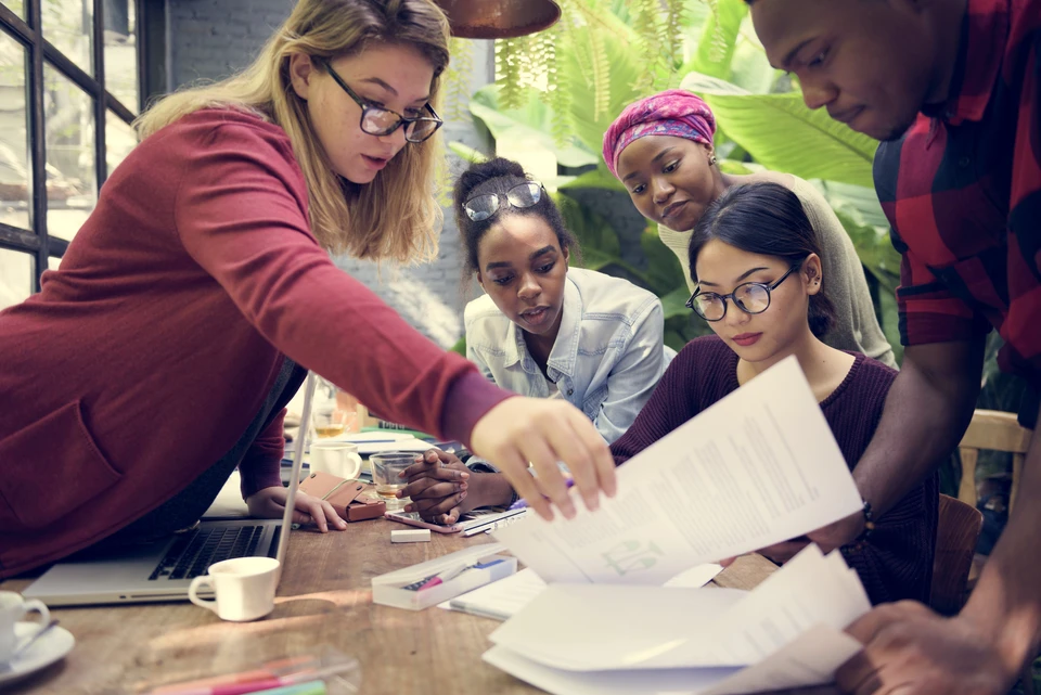 Une femme blonde en blazer rouge présente des documents à quatre collègues réunis autour d'une table de travail dans un bureau moderne avec végétation
